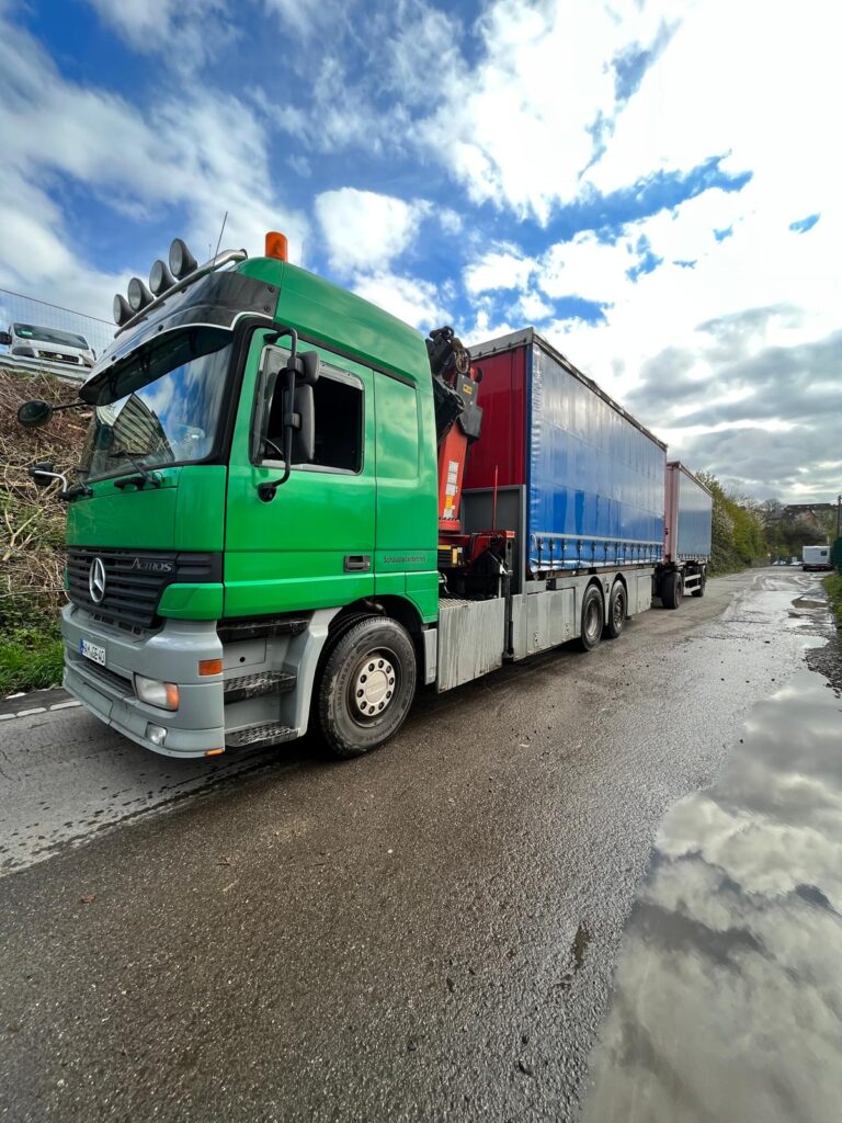 Gruener Mercedes Actros Lastwagen mit Ladekran und blauer Plane am Strassenrand