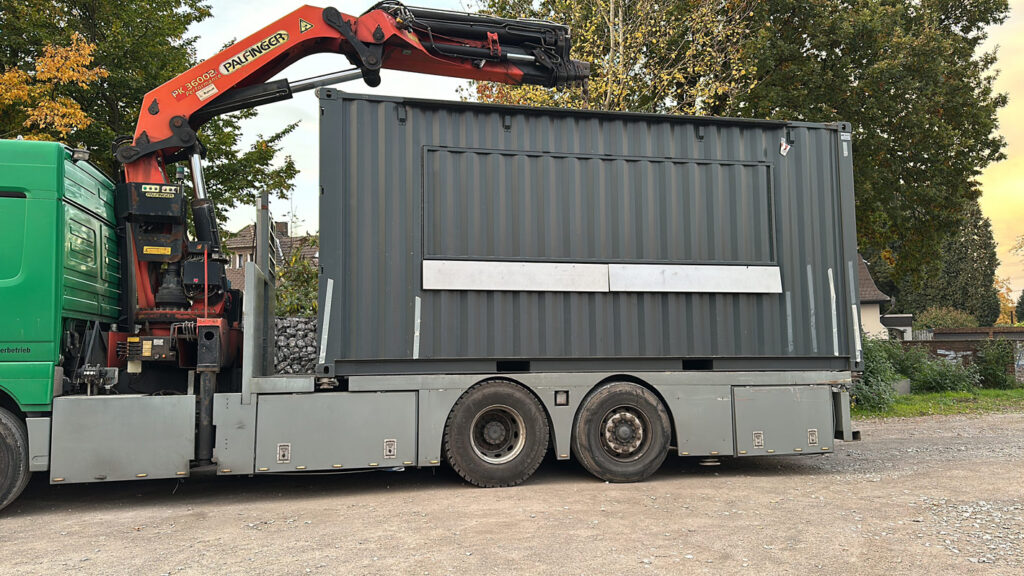 Gruener Lastwagen mit rotem Palfinger Kran transportiert grauen Container